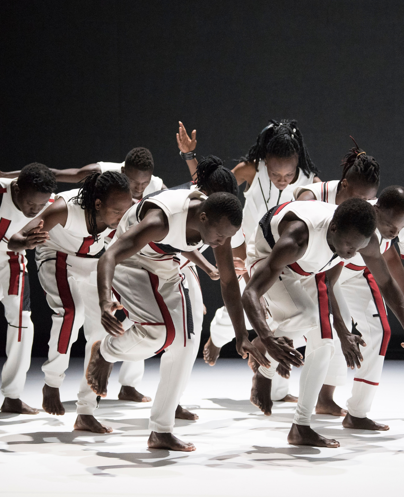 9 African dancers dressed in white with red and black trim move rhythmically in unison