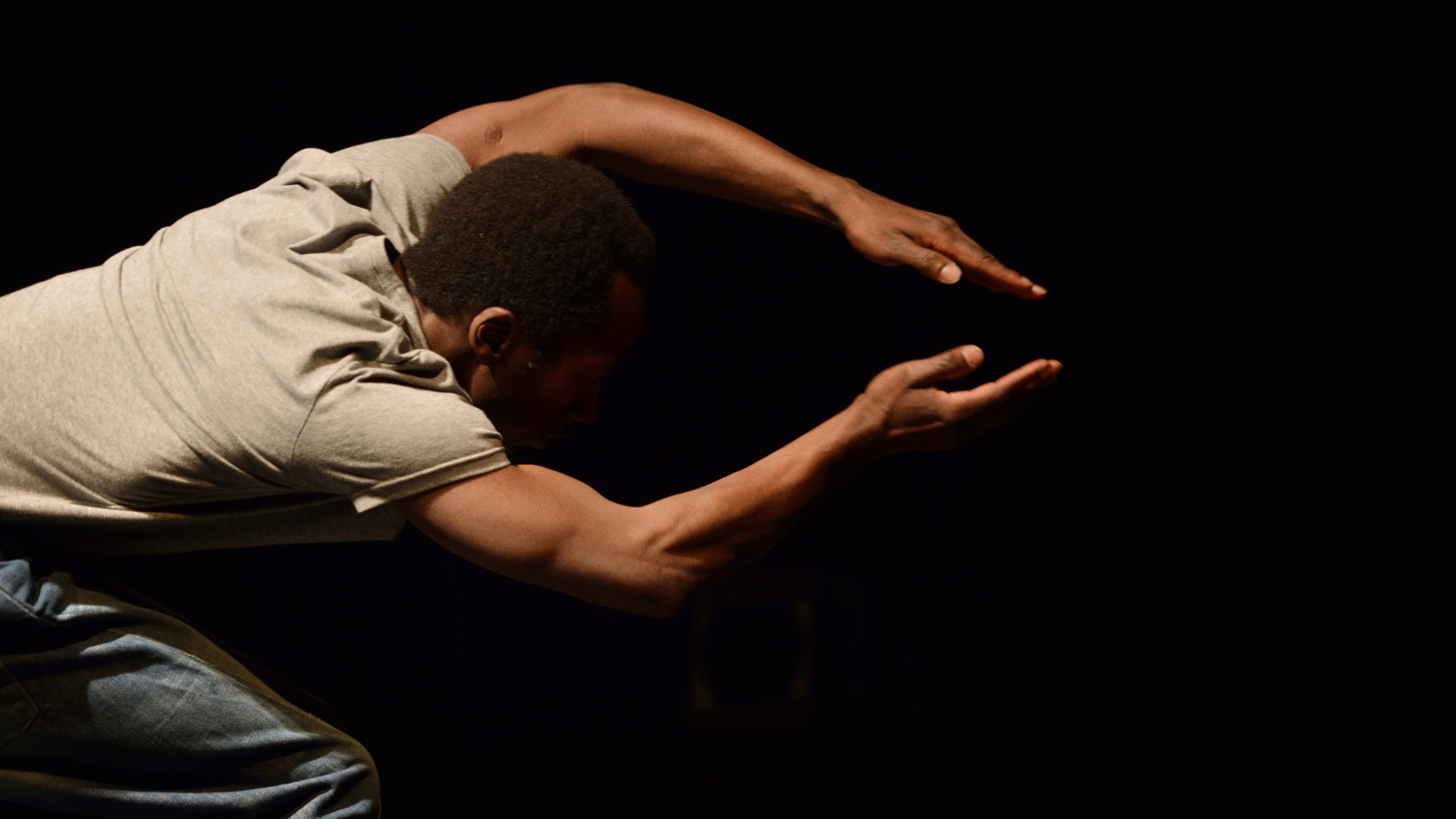 Close up of a dancer with arms forming an arch over his head. He is in a dimly lit space and wears a neutral coloured tshirt.