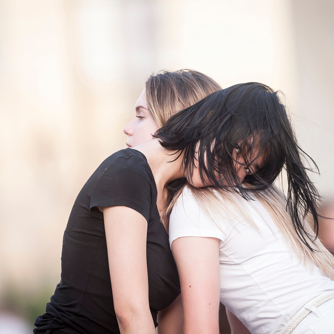 Two dancers rest their heads on each others' shoulders in an outdoor performance