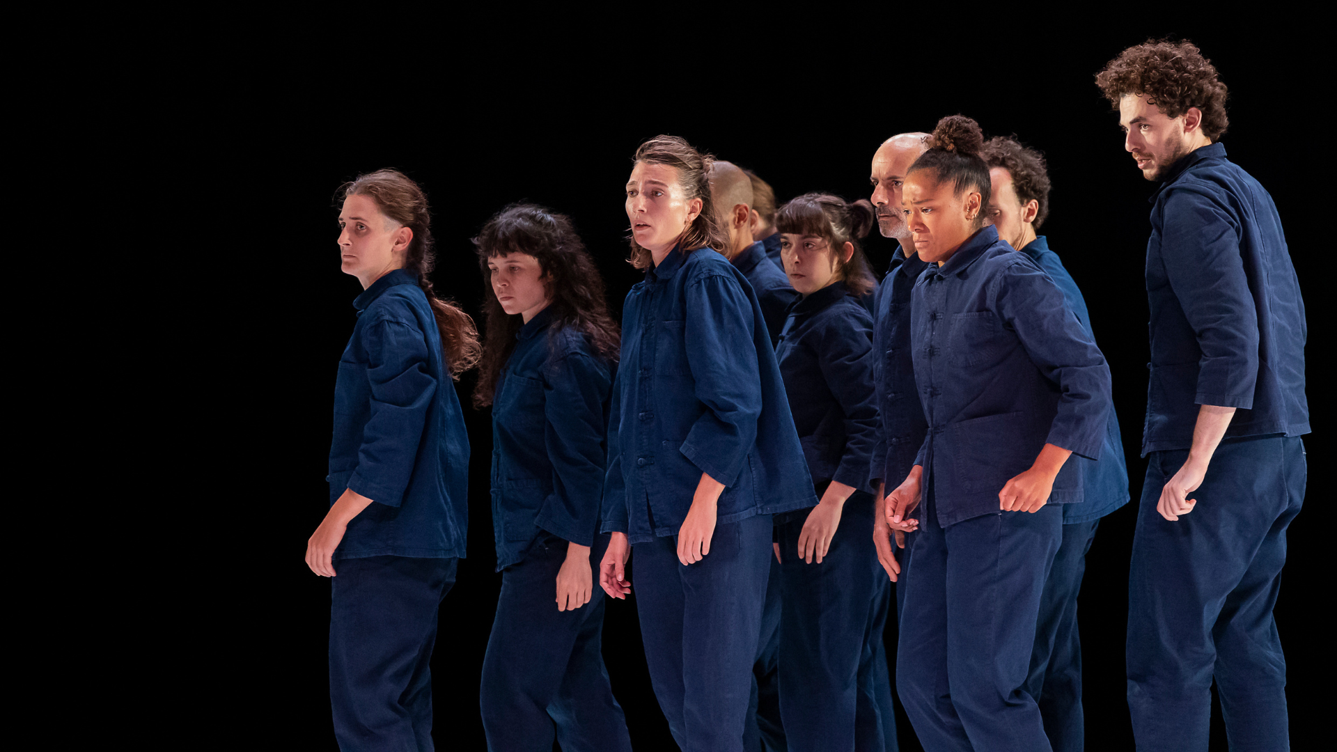 Group of performers in blue jump suits stand together all facing the same direction on a dimly lit stage