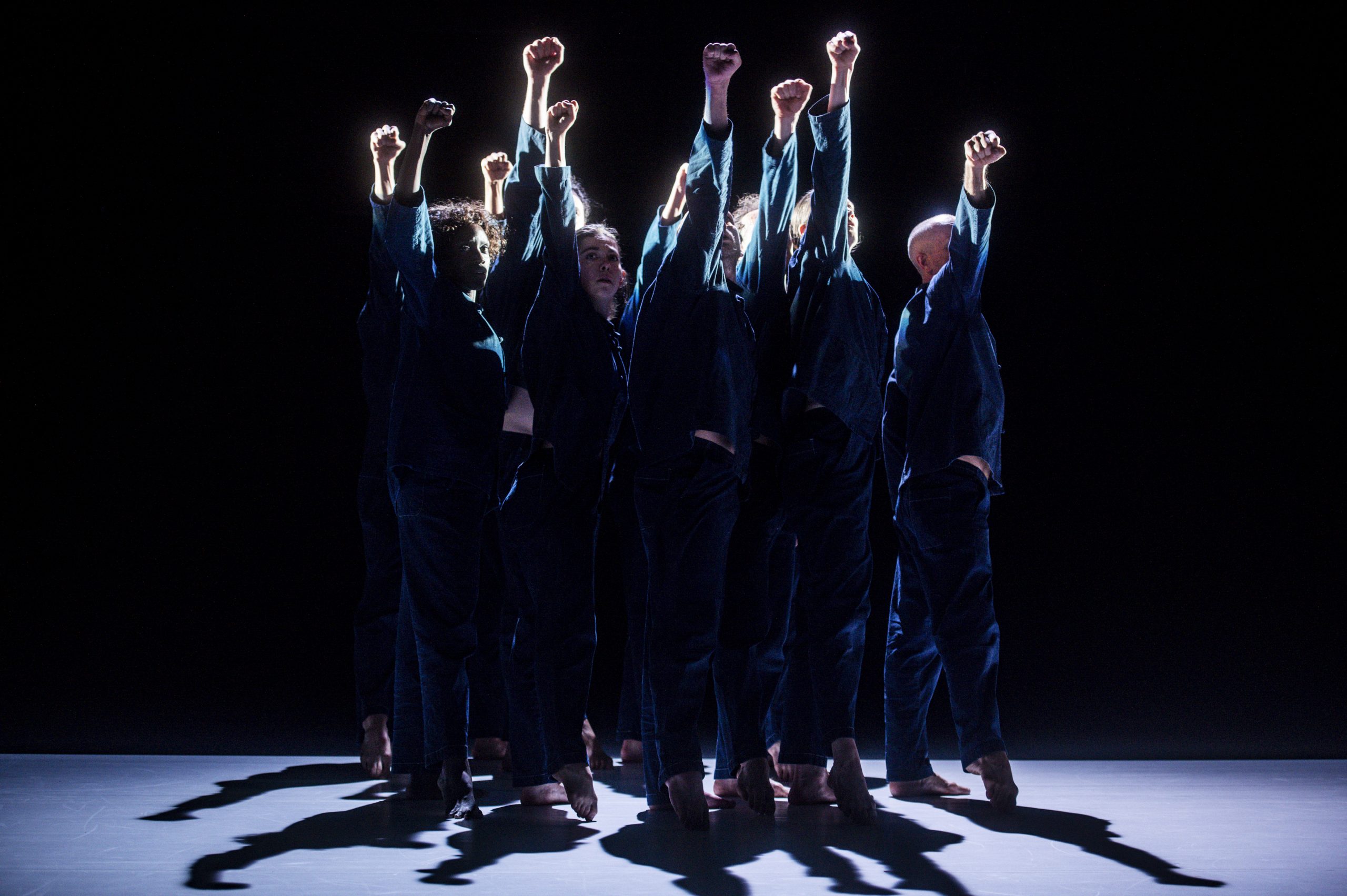 A group of dancers in navy jumpsuits form a close huddle on a dark stage and each lift their right arms into the air holding a tight fist.