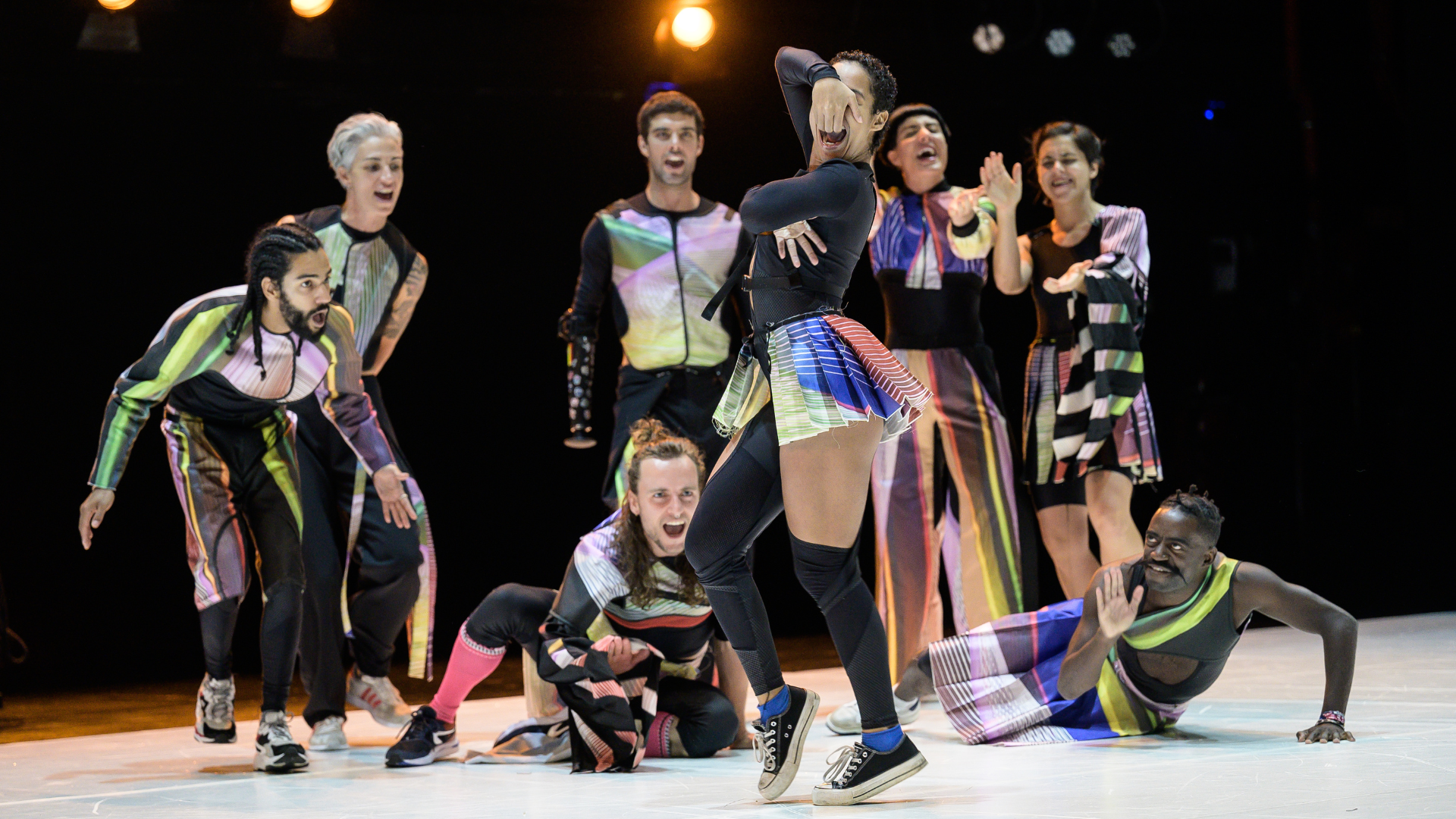 A group of dancers in luminous clothing gather together and cheer on one performer as she dances