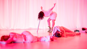 Bathed in bright pink light, dancers are lying on the floor while one poses with her head resting on a baseball bat