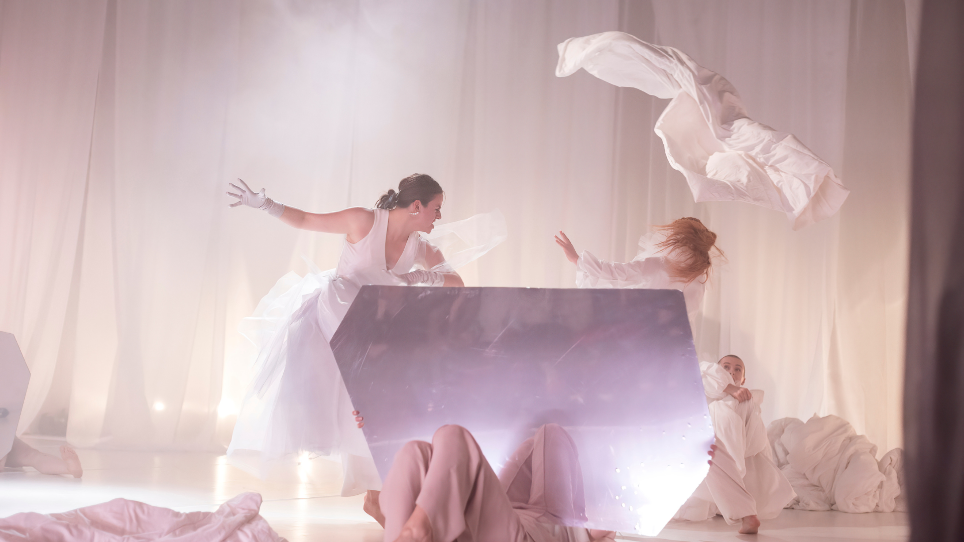 White fabric floats in the air as a dancer moves and gestures to the sky. She is also dressed in a flowing white costume.