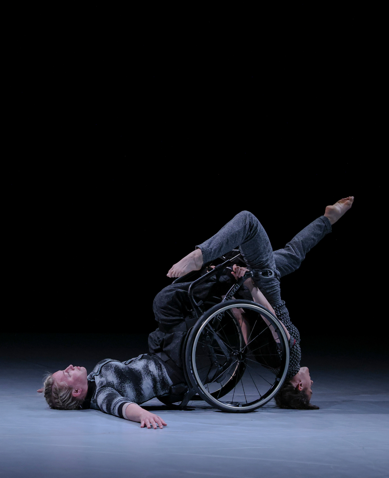Two artists perform on a bare stage using a wheelchair to balance on and lean against as one does a headstand and the other lies with her back on the floor.